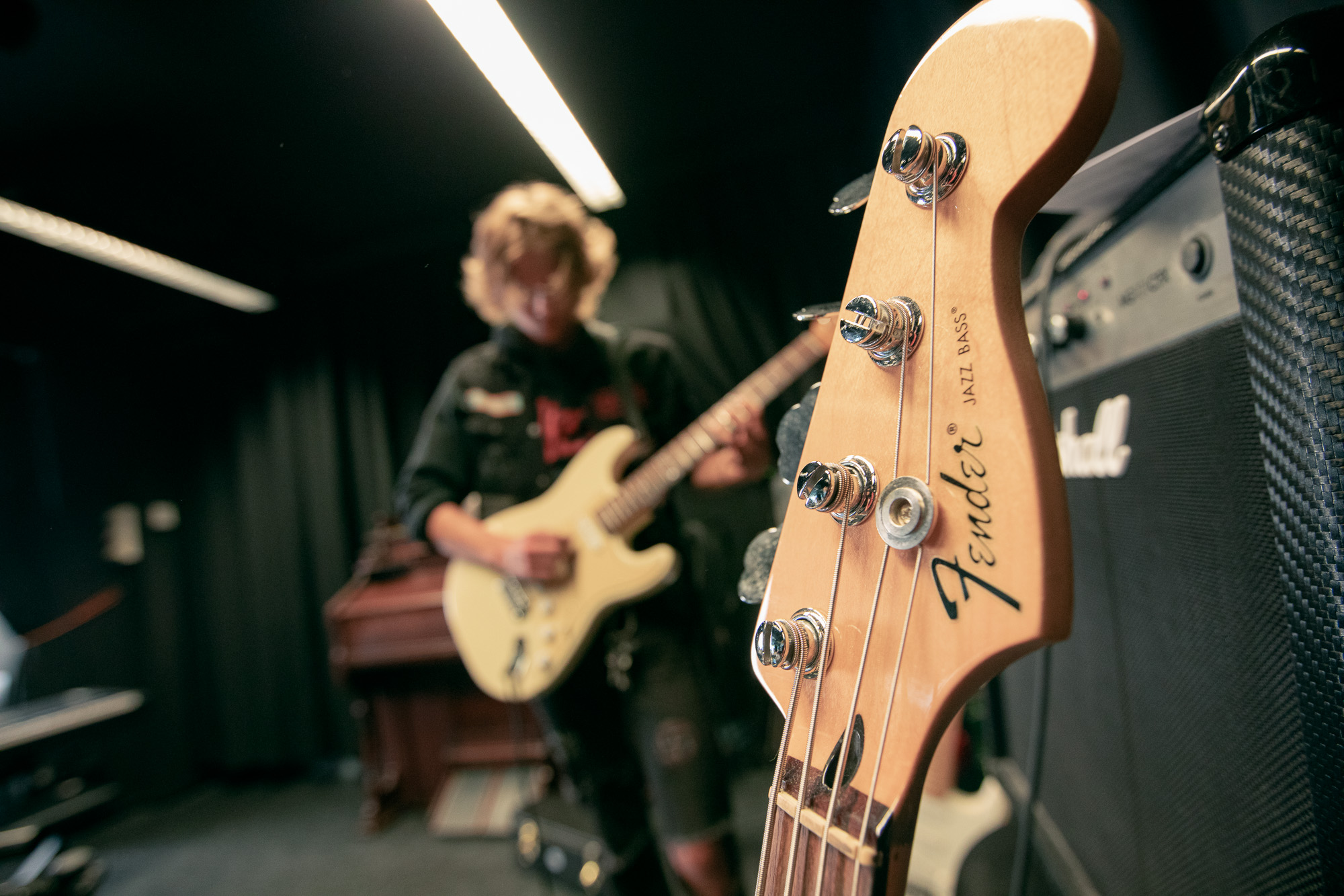 Fender-basson lapa ja virityskoneisto sekä Marshallin kaiutin, taustalla nuori soittaa sähkökitaraa.